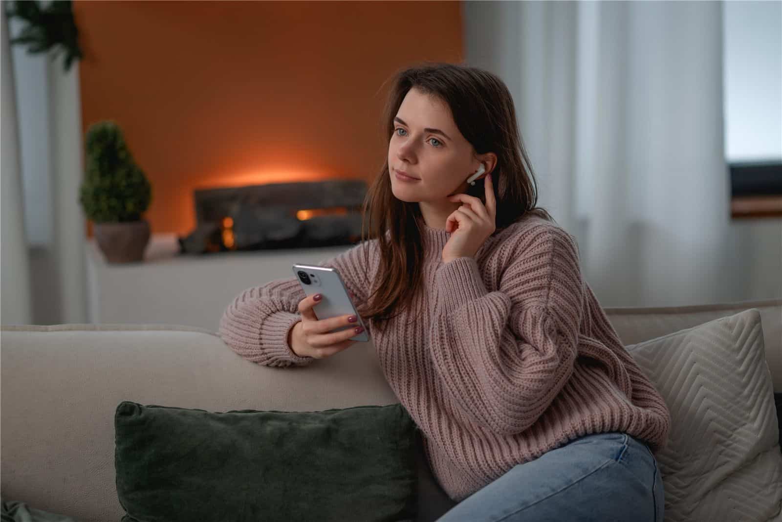 Junge Frau mit Kopfhörern und Smartphone auf dem Sofa, warmes Licht